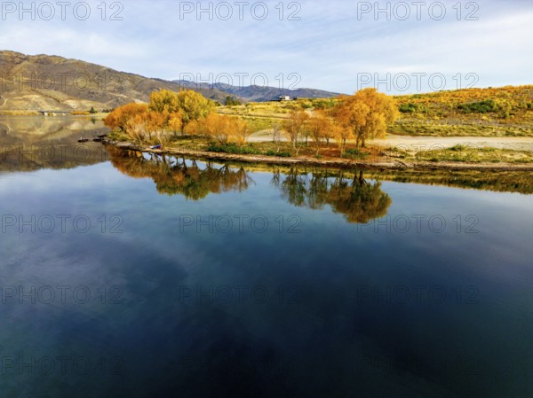 Aerial view of a tranquil lake in Queenstown during autumn, showcasing vibrant foliage and clear reflections on the water surface. Captured by a drone for a stunning aerial perspective