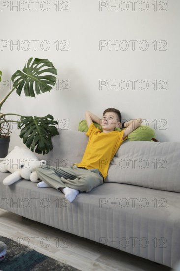 A young boy in a yellow shirt relaxes on a gray couch, leaning against green avocado plush toy and a fluffy toy. A large plant adds a touch of greenery to the cozy scene