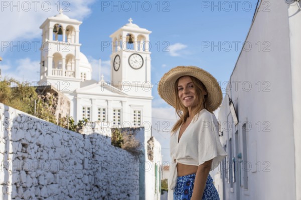A smiling woman in a straw hat enjoys a sunny day exploring the charming village of Trypiti, Milos Island, Greece, with iconic white architecture and blue skies