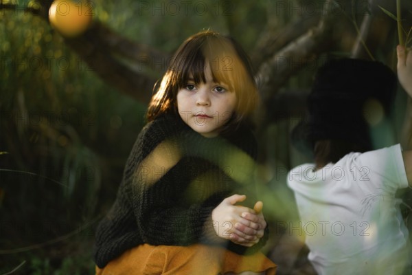 A young child with thoughtful expression sits in a forest setting. Warm sunlight filters through leaves, adding a dreamy feel to the serene natural scene