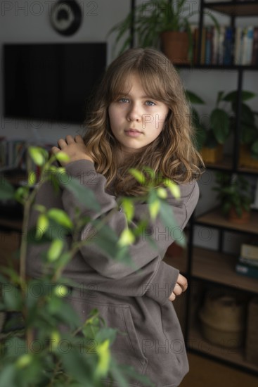 A teenage girl stands thoughtfully in a cozy home, surrounded by houseplants and a bookshelf. Her casual attire and calm demeanor create a serene, welcoming ambiance