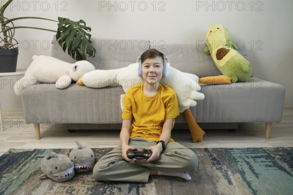 A cheerful boy in a yellow shirt and headphones plays video games, seated in a cozy room with plush toys and a monstera plant on a comfy sofa