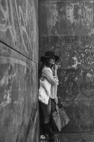 A woman wearing a stylish hat and casual attire leans against a textured, urban wall, exuding modernity and elegance. Her thoughtful expression adds depth to the scene
