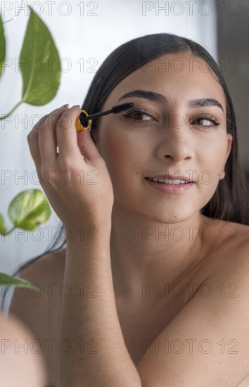 A woman smiling as she applies mascara in a brightly lit room. The presence of a vibrant green plant in the background adds a natural touch to the scene