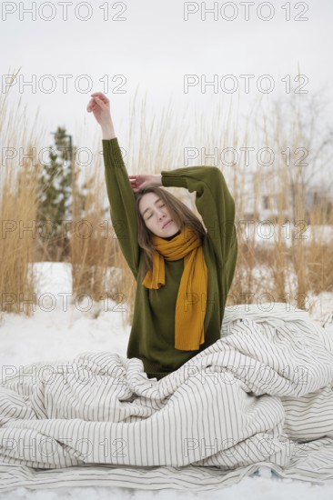 A woman with a serene expression stretches in a snowy park, wearing a green sweater and yellow scarf, surrounded by tall grasses and wrapped in a striped blanket