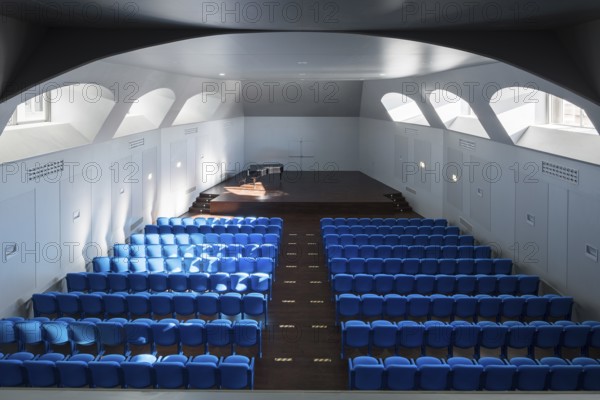 A spacious auditorium with rows of vibrant blue seats facing a stage. Natural light streams through upper windows, creating a serene atmosphere for performances or music events