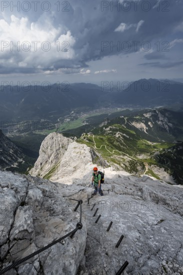 Mountaineer with helmet climbing on the rock in a secured climb, on a narrow rocky ridge, in steep rocky mountain landscape, view of Garmisch Partenkirchen, descent from the Alpsitze via the via ferrata, Alpspitz Ferrata, Wetterstein range, Bavaria, Germany