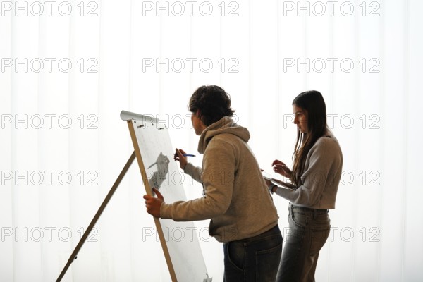 Two teenagers, a non-binary individual and a female, participate in an writing activity, o a whiteboard easel in a brightly lit classroom environment. Their engagement illustrates a vibrant example of learning in an educational setting