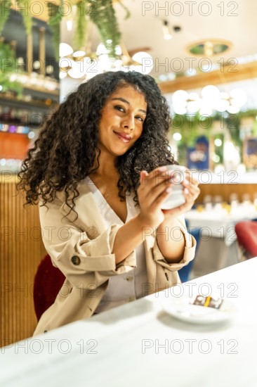 Vertical portrait of a latin beauty woman enjoying warm coffee sitting in a modern cafeteria smiling at camera