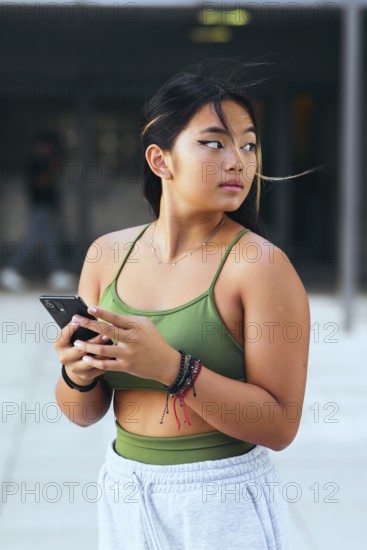 A teenage girl holds a smartphone while standing outdoors. Dressed in a green top and light pants, she gazes thoughtfully, embodying modern youth culture and connectivity
