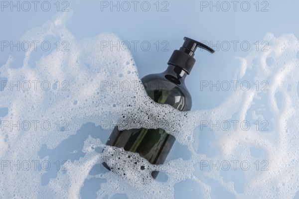 A sleek black shower gel bottle lies amidst generous white foam on a soothing blue background. Perfect for beauty, skincare, and product marketing visuals