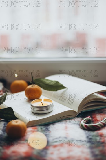 Warm and inviting Christmas scene featuring a lit candle placed on an open book, surrounded by fresh tangerines and a candy cane, offering a sense of holiday cheer and relaxation