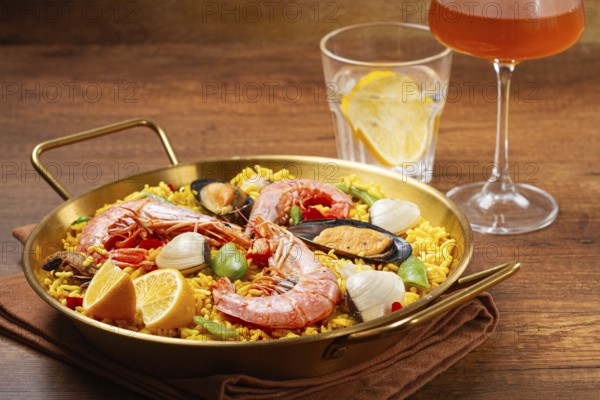 A large dish of seafood paella features shrimp, mussels, and other ingredients. A glass with a drink and slice of lemon is nearby on the wooden table. The meal is ready to eat