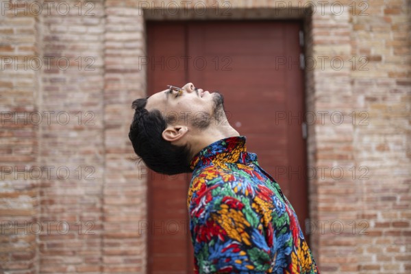 A fashionable cuban man wearing a colorful tropical shirt leans back with his head tilted and glasses slipping down his nose. The artistic pose and brick background create a modern, expressive vibe