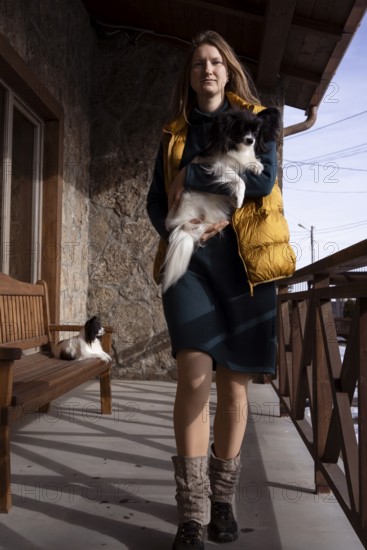 A woman stands on a rustic porch holding her purebred papillon dog. Natural light illuminates the charming scene, capturing the bond between owner and pet