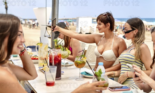 Friends gather around a beach bar, savoring colorful cocktails and laughing together. Some take selfies while enjoying the vibrant summer atmosphere on a sunny day by the ocean