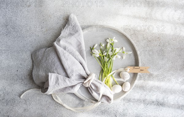 Top view of a decorative spring setup featuring a cluster of fresh snowdrops in a ceramic plate, accompanied by a napkin, eggs, and a Thank you note on a textured background