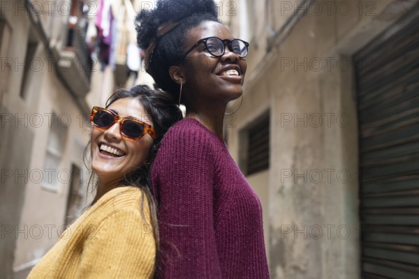 Two friends smiling joyfully, standing back-to-back in a narrow street. One wears sunglasses and a mustard sweater, the other glasses and a burgundy sweater, enjoying the sunny day