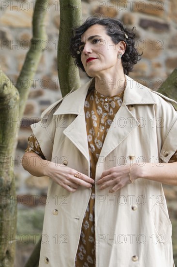 A fashion woman in a beige trench coat and patterned dress stands gracefully outdoors. The stone wall and tree add a rustic charm, highlighting her elegant style