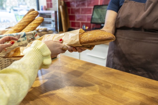 Customer hands collecting a freshly baked baguette wrapped in paper from a baker behind a wooden counter, symbolizing freshness, local shopping, and daily bakery purchases