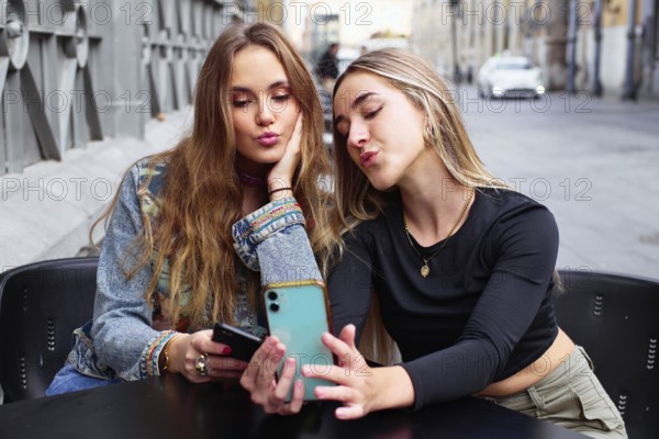 Two teenage girls sit together, using their cell phones and sharing a fun moment. They are outdoors, smiling and enjoying each other's company in a casual setting