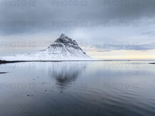 A peaceful winter scene of Kirkjufell Mountain in Iceland, covered in snow. The mountain reflects in the calm water, under an overcast sky at twilight