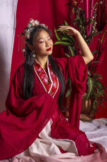 A woman in a red robe sits on a bed with a potted plant behind her. She is wearing a flower headband and has red lipstick on. Concept of elegance and cultural appreciation