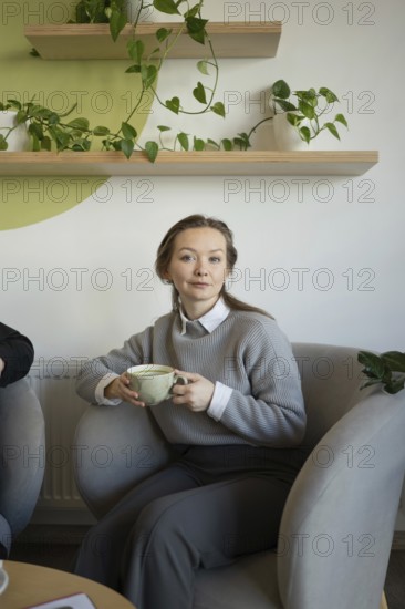 A woman sits comfortably in a cafe, sipping coffee. She is surrounded by green plants, creating a relaxing and inviting atmosphere. The serene scene embodies comfort