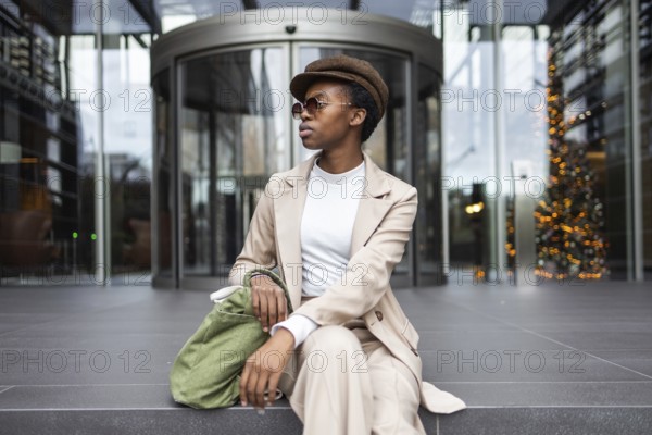 A fashionable black woman in a beige suit, sitting outside a modern building with glass doors She exudes confidence and style in an urban setting