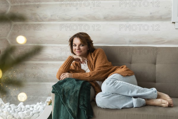 A woman in a cozy brown cardigan and blue pants lounges on a gray sofa, looking at the camera amidst Christmas decorations. Her warm smile adds to the festive ambiance reflected by the twinkling lights and a green blanket beside her