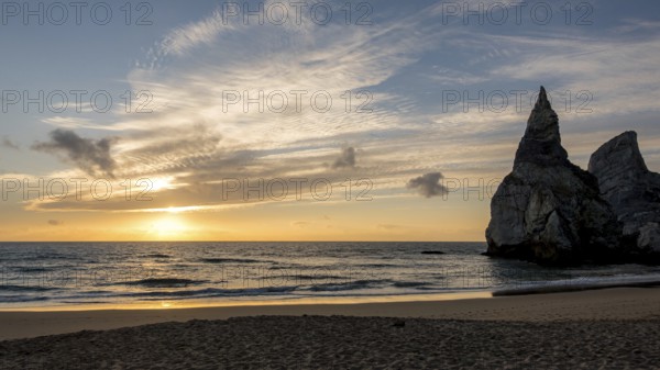 A stunning sunset over a tranquil ocean with majestic rock formations on the shoreline of bear beach, Portugal. The sky is painted with warm hues, reflecting on the water, creating a peaceful scene