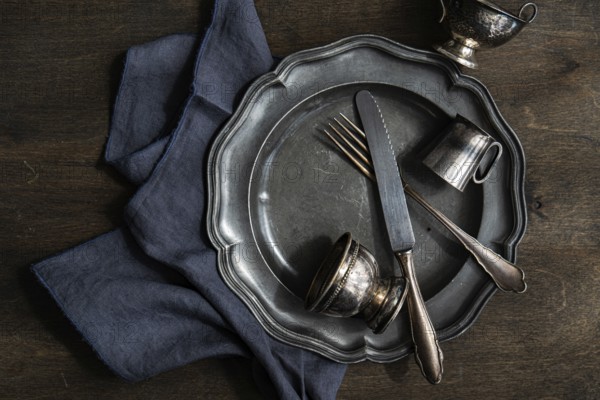 A collection of vintage silver plate, cutlery, and cups arranged on a dark wooden surface with a blue cloth. The tarnished metal adds a rustic, antique touch