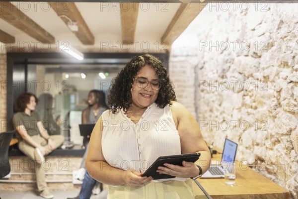 Multiracial coworkers collaborate in a modern office. A woman uses a tablet while others engage in conversation, showcasing a diverse and inclusive work environment