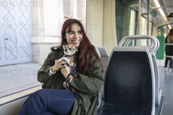 A cheerful woman sits on a tram seat holding her small chihuahua, warmly wrapped in a coat. The tram modern interior creates a cozy setting for their journey