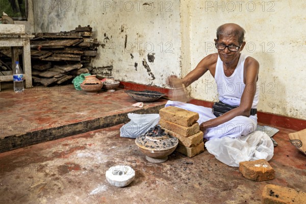 Man sitting smiling in a workshop with bricks and braziers, a man burning sapphires in a small oven in the town of Ratnapura in Sri Lanka