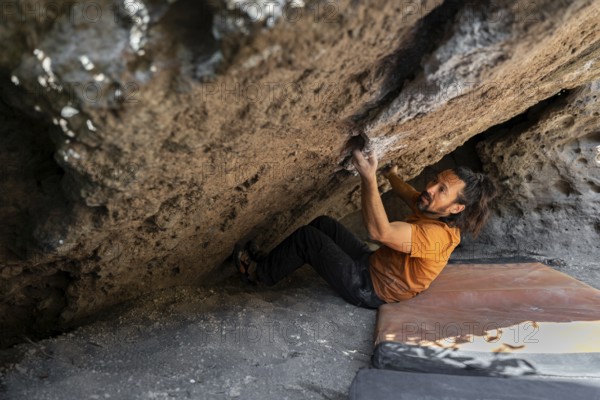A climber in an orange shirt tackles a challenging boulder face in the wilderness, gripping rocks with determination. The scene is set against a backdrop of rugged natural beauty