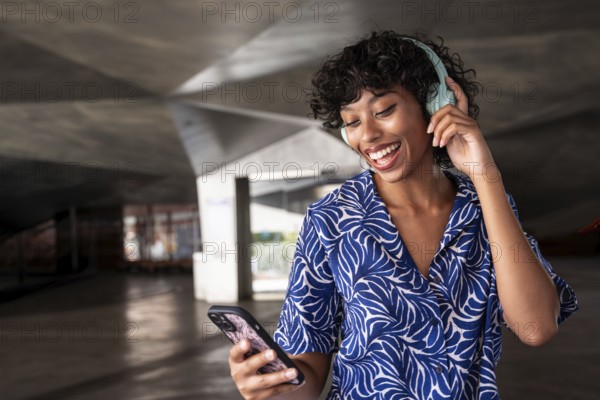 A joyful woman enjoys music through her headphones while holding a smartphone, standing in a modern architectural space. She smiles brightly, expressing contentment