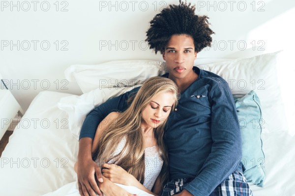 A serene moment between a couple lounging comfortably in bed. The woman rests her head on the man's chest, exuding warmth and mutual affection in a cozy setting