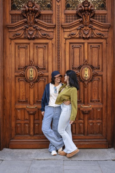 A lesbian couple smiling and embracing in front of an ornate wooden door, exuding love and happiness in an urban environment. Casual and warm atmosphere captured in the city