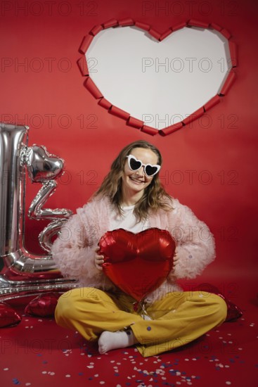 A young woman smiles joyously while holding a red heart-shaped balloon. She wears heart-shaped sunglasses and sits against a vibrant red background with Valentine's Day decorations