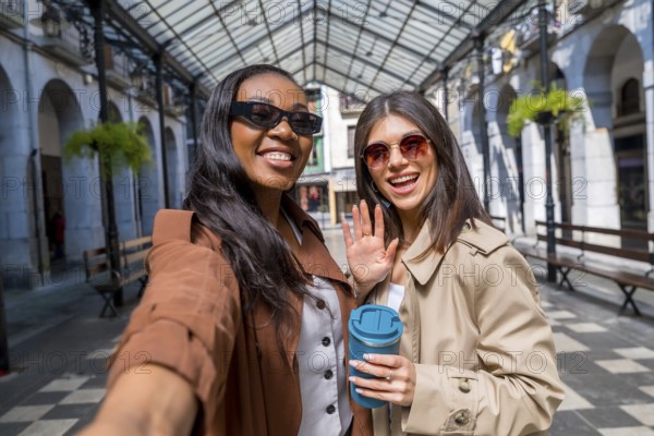 Two smiling businesswomen taking a selfie and waving at the camera in a city center gallery while holding a reusable coffee cup