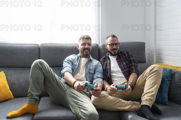 A joyful gay couple relaxes on a couch, engaging in a lively video game. Their cheerful expressions highlight a moment of leisure and connection, capturing modern home life