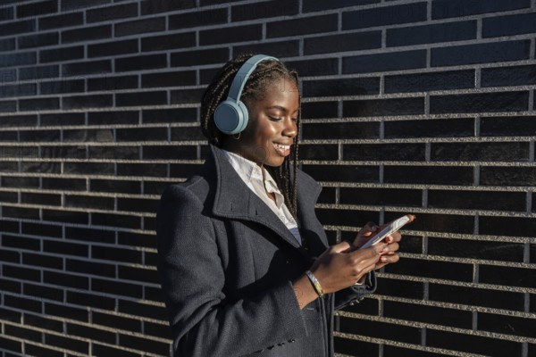 Side view of a young African American businesswoman with braids is seen enjoying a break, smiling while using her smartphone and listening to music through headphones outdoors