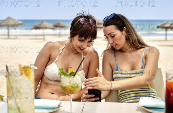 Two women sit together at a beach side table enjoying refreshing summer cocktails. Engaged in conversation, they share moments while looking at their phones, surrounded by sun and sand