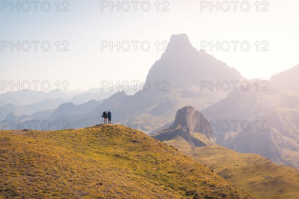 Two hikers stand on a grassy hill, gazing at the breathtaking mountain range in the Pyrenees. The vast landscape and early light create a serene and inspiring scene in summer