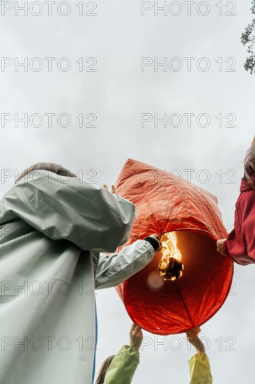 From below unrecognizable people in Taiwan lifting a glowing red Chinese lantern ready for flight. Participants, clad in raincoats, prepare this lantern, often inscribed with wishes for health, happiness, and prosperity
