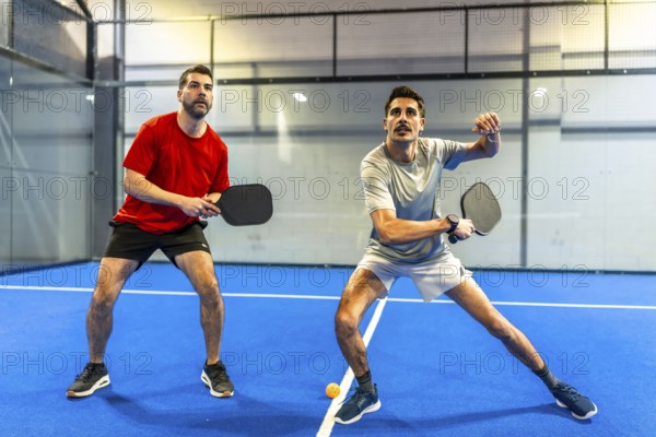 Two active men engaging in a pickleball match on a blue indoor court, concentrating on hitting the ball with their paddles during an athletic competition