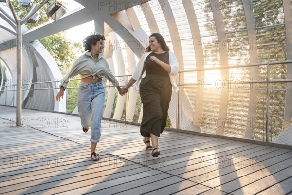 Lesbian couple joyfully holds hands while running together on a modern walkway, surrounded by sunlight and greenery. A moment of freedom and happiness captured