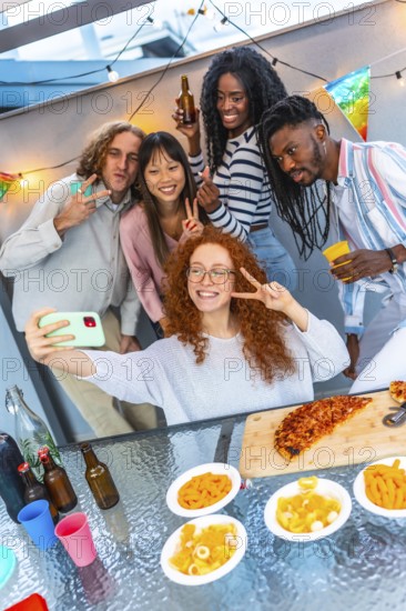 Vertical photo of a group of multi-ethnic friends gesturing success while taking selfie during party in a rooftop at evening