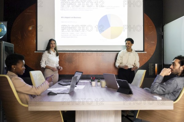 A diverse team engages in a modern office meeting. Two presenters stand by a screen displaying a pie chart, while colleagues sit attentively with laptops on the table